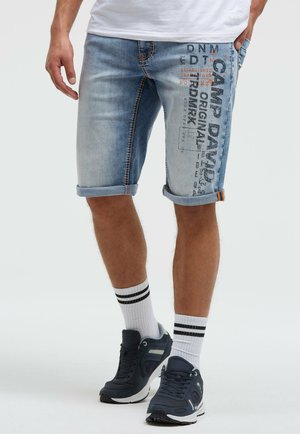Mann trägt hellblaue, hochgekrempelte Jeansshorts mit bedrucktem Text, weiße Socken mit schwarzen Streifen und schwarze Sportschuhe.