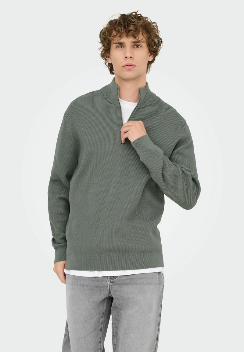 Groen geribbeld trui met een halve rits kraag, lange mouwen en een relaxte pasvorm, gedragen over een witte t-shirt, gecombineerd met lichtgrijze jeans.