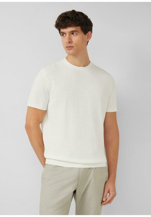 Mężczyzna z kręconymi ciemnymi włosami, ubrany w teksturowany, biały, krótki sweter z krótkim rękawem oraz jasnoszare spodnie, stojący na jednolitym szarym tle.