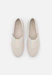 Sapatos beje tipo slip-on feitos de lona texturizada, com pormenores elásticos nas laterais e um design de ponta arredondada suave. Sola em borracha branca.