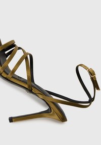 Oliven grønn høyhælte sandal med sorte stropper, spiss tå og en slank stiletto hæl. Har en ankelstropp med en gullfarget spenne.