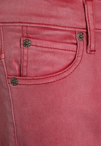 Punainen denimpuku, jossa on sileä pinta, etutasku ja metallinieteet. Ompeleet ovat sävyn mukaan ja pinta näyttää hieman haalistuneelta.