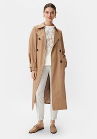 Beige trenchcoat met dubbelzijdige knopen, voorzakken en brede revers, gedragen over een wit graphic t-shirt en smalle witte broek.