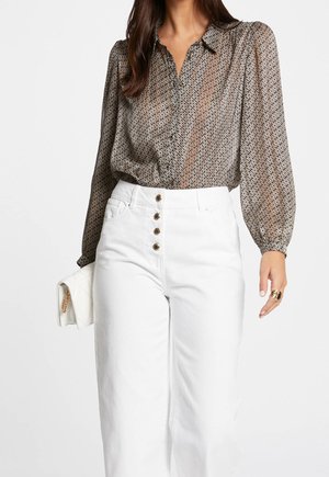 Femme portant un chemisier à manches longues à motifs rentré dans un pantalon blanc taille haute avec des boutons dorés, tenant une pochette blanche matelassée.