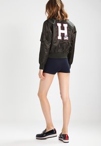 Svart satin bomberjacka med vit och röd "H" brodyr på baksidan, åtsittande manschetter och djupa fickor; matchad med marinblå shorts och glänsande skor.