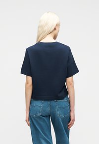 Ξανθιά γυναίκα που φοράει απλό μπλε σκούρο κοντομάνικο T-shirt και μπλε τζιν, γυρισμένη προς τα πίσω σε ένα απλό φωτεινό φόντο.