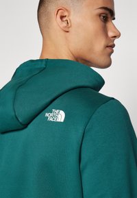 Teal színű kapucnis pulóver a The North Face-től, fehér logóval a hátán. Sima anyagból készült, struktúrált kapucnival. Közeli nézet a nyakrészről.