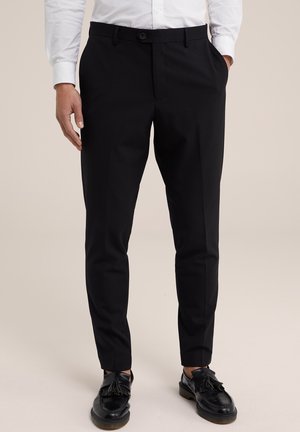 Homme portant un pantalon de costume noir slim, une chemise blanche rentrée, et des mocassins noirs à pampilles, debout les mains partiellement dans les poches.