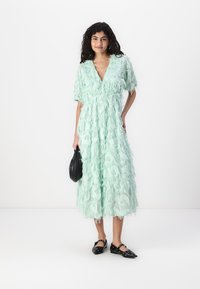 Light mint green dress featuring layered, textured ruffles, short sleeves, and a V-neckline, paired with a black bag and black flats.
