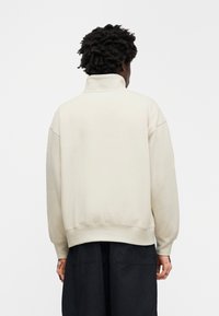 Μπεζ μπλούζα με ψηλό γιακά, άνετη γραμμή, χαμηλωμένους ώμους και ριμπ μανσέτες. Λεια υφή και μινιμαλιστικός σχεδιασμός.