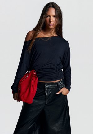 Femme aux longs cheveux bruns portant un haut noir décolleté aux épaules et un pantalon en cuir noir, tenant un grand sac à main rouge sur son côté gauche.