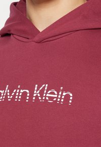 Bordowy hoodie z dużym białym logo "Calvin Klein" w paski. Wykonany z miękkiego materiału w klasycznym kroju z kapturem.