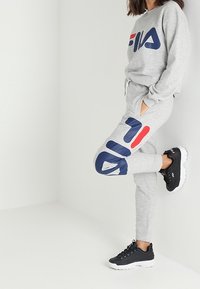 Gray sweatshirt and joggers featuring navy and red logo design, made of soft fabric. Black athletic shoes with white soles complete the look.