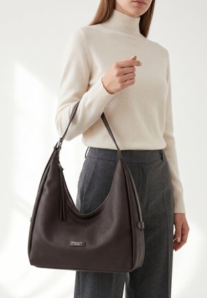 Femme portant un col roulé crème et un pantalon gris, tenant un grand sac à main marron foncé texturé avec une bandoulière et une tirette de fermeture éclair.