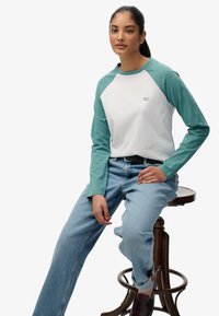 Langarmshirt met een witte body en teal mouwen, gemaakt van zacht materiaal. Heeft een klein logo op de borst. Gemaakt in combinatie met lichtblauwe jeans.