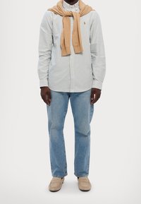 Camicia a righe blu e bianca, con un maglione beige annodato intorno al collo, jeans azzurri e mocassini beige.