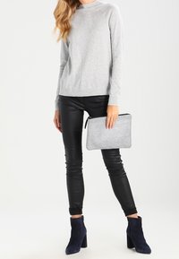 Frau in grauem Pullover und schwarzen Lederhosen, die eine silberne Clutch hält und navyblaue Absatzstiefel trägt.