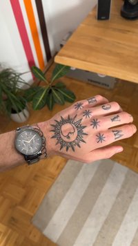 Hõbedane käekell käel, millel on keerukad mustad tätoveeringud, sealhulgas päikese, kuu ja tähe motiivid, puidust laua taustal.