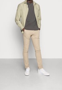 Světlá zelená košile na knoflíky, šedé tričko, béžové přiléhavé kalhoty, bílé tenisky. Design je neformální s hladkou texturou a minimálními akcenty.