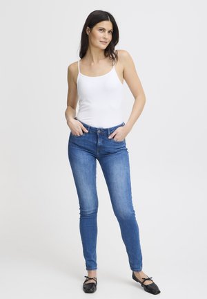 Femme aux cheveux noirs portant un débardeur blanc, un jean skinny bleu et des ballerines noires avec des nœuds, debout les mains dans les poches.