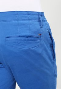 Modré bavlněné kalhoty s hladkou texturou, které mají zadní kapsu a malou logo aplikaci v červené, bílé a modré barvě na pase.