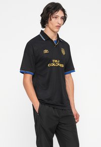 Νεαρός άνδρας με μαύρα σγουρά μαλλιά, φορώντας μαύρη αθλητική ζακέτα με χρυσά γράμματα "True Colors" και λογότυπα, με τα χέρια στις τσέπες, στέκεται μπροστά σε λευκό φόντο.