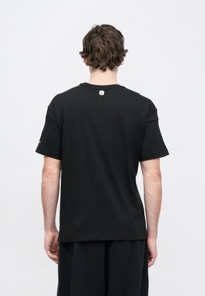 Mężczyzna o krótkich kręconych włosach, ubrany w zwykły czarny T-shirt z krótkim rękawem, zwrócony tyłem na jasnoszarym tle.