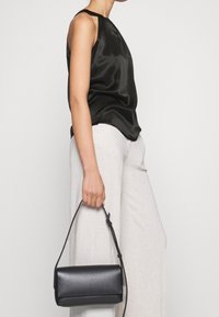 Černý saténový top bez rukávů s kulatým výstřihem, doplněný světle zbarvenými lněnými kalhotami s širokými nohavicemi a obdélníkovou černou kabelkou.