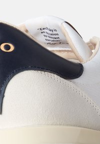 Sneaker bianco con tacco in suede beige testurizzato, parte superiore blu scuro e linguetta in tessuto intrecciato che presenta una citazione in testo nero all'interno.