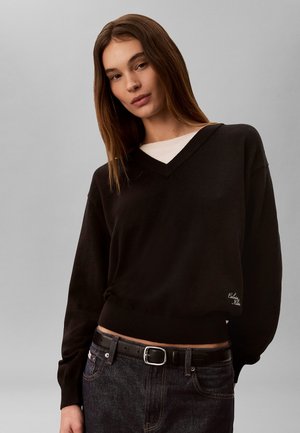 Jeune femme aux cheveux bruns portant un pull noir à col en V par-dessus un haut blanc et un jean foncé avec une ceinture noire sur un fond gris uni.
