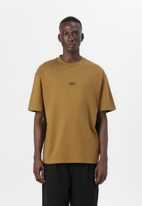 Brun bomulls t-skjorte med rund hals, korte ermer og et lite svart logo-patch på brystet. Avslappet, boksformet passform.