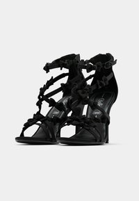 Melni augstpapēžu sandales ar vairākiem siksniņām, rotātas ar mazām lokām un potīšu sprādzēm uz baltas fona.