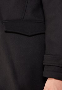 Cappotto nero con finitura testurizzata, tasca a falda sul petto e dettagli sui polsini con tre cuciture orizzontali.