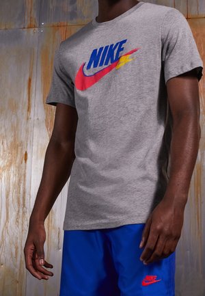 Szary bawełniany T-shirt z dużym, kolorowym logo Nike w kolorach niebieskim, czerwonym i żółtym. Model ubrany w niebieskie szorty, prezentujący sportową sylwetkę.