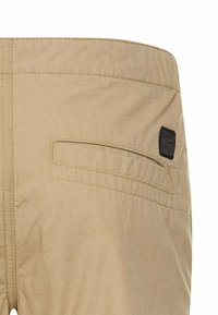 Beige Hosen mit einer glatten Textur, elastischem Bund und einer einzelnen Gesäßtasche mit einem schwarzen Akzentlabel. Einfaches Design und neutrale Farbe.