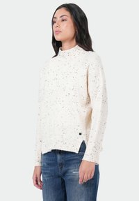 Crème kleurige gebreide trui met een hoge kraag, gestructureerde textuur, geribbelde manchetten en zijsplitten, gecombineerd met blauwe distressed jeans.