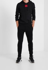 Sudadera negra con un parche de logo rojo, bolsillo canguro y capucha ajustable, combinada con pantalones jogger negros ajustados y zapatillas deportivas negras.