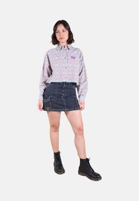 Chemise à manches longues bleue et rose avec un motif en crochet, portée avec une mini-jupe en denim foncé dotée de poches et d'un patch logo. Bottes noires à lacets.