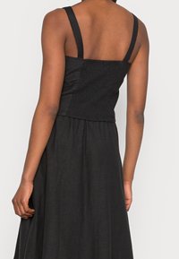 Robe noire sans manches avec un corsage ajusté mettant en valeur des détails froncés et une jupe évasée. Le tissu a une apparence douce et texturée.