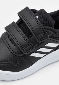 Zapatillas deportivas sintéticas negras con dos correas ajustables de velcro, rayas blancas de acento, forro de malla negra texturizada y suela de goma blanca.