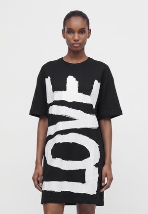 Giovane donna che indossa un vestito T-shirt nero oversize con grandi lettere bianche a pennellate sul davanti, in piedi su uno sfondo bianco uniforme.
