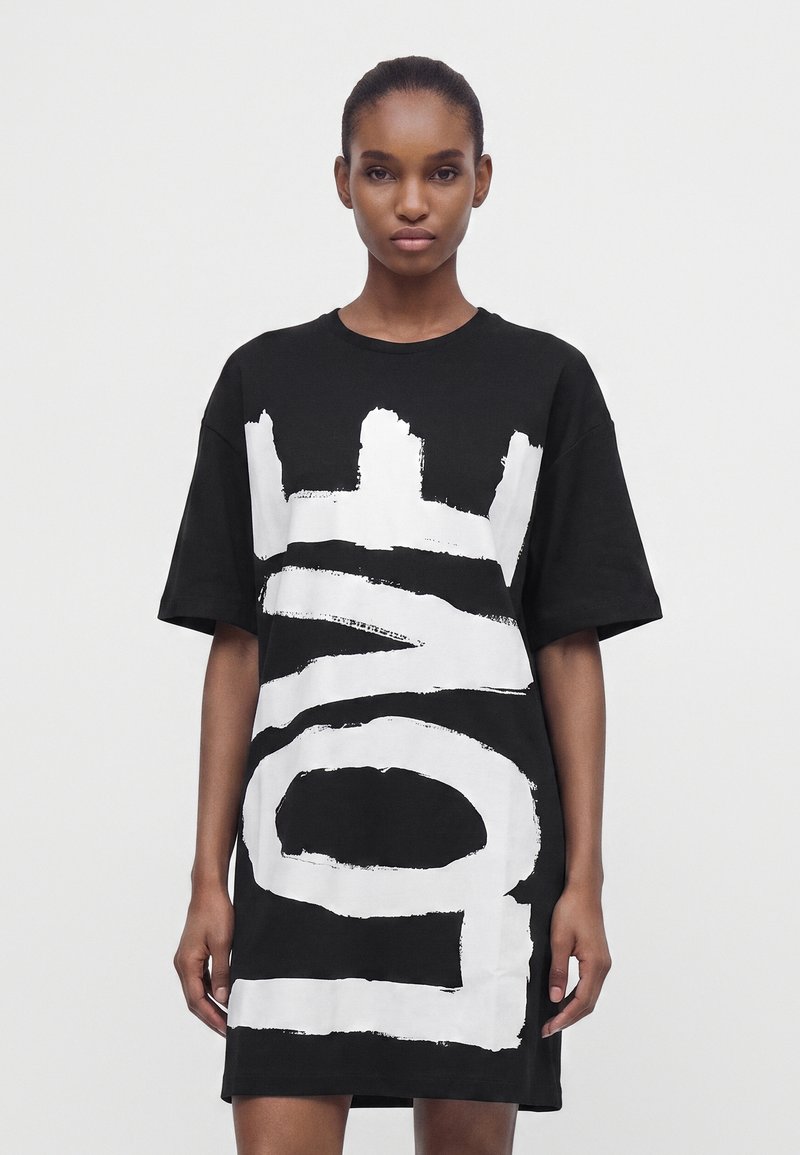 Νεαρή γυναίκα που φοράει μαύρο oversized φόρεμα T-shirt με μεγάλα λευκά γράμματα σε πινελιές μπροστά, στέκεται μπροστά σε απλό λευκό φόντο.