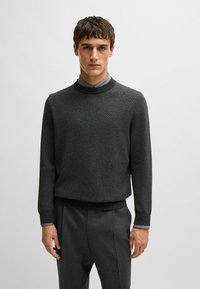 Maglione grigio scuro a maglia con scollo rotondo, motivo testurizzato e polsini a costine, indossato sopra una camicia con colletto. Abbinato a pantaloni grigi.