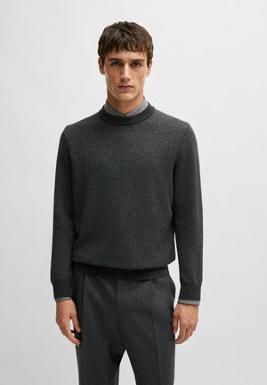 Dunkelgrauer gestrickter Pullover mit rundem Halsausschnitt, strukturiertem Muster und gerippten Bündchen, getragen über einem Hemd mit Kragen. Kombiniert mit grauen Hosen.