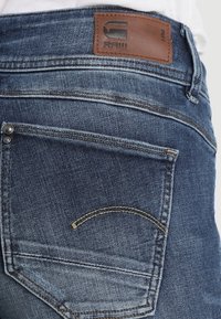 Mörkblå denimjeans med en läderlapp märkt "RAW" vid midjan, med detaljerade sömmar och en bakficka med böjd söm.