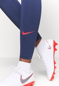 Personne portant des leggings bleu marine avec un logo Nike rouge et des chaussures de football Nike blanches avec semelles et accents rouges, debout sur une surface blanche.