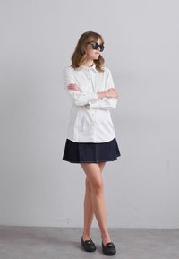 Witte blouse met lange mouwen, gecombineerd met een donkerdenim A-lijn rok. Zwarte zonnebril en instappers maken de look af.
