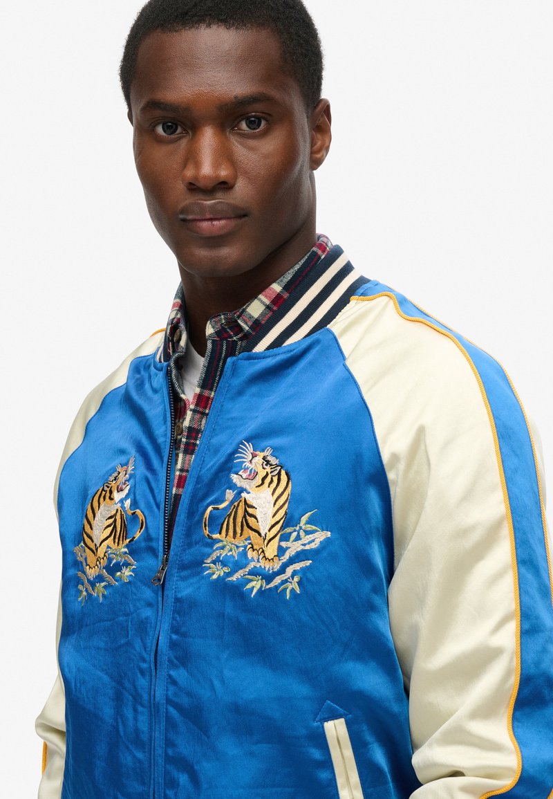 Homme portant une veste en satin bleu et crème avec des tigres brodés sur la poitrine et une chemise à carreaux en dessous, faisant face à l'objectif.