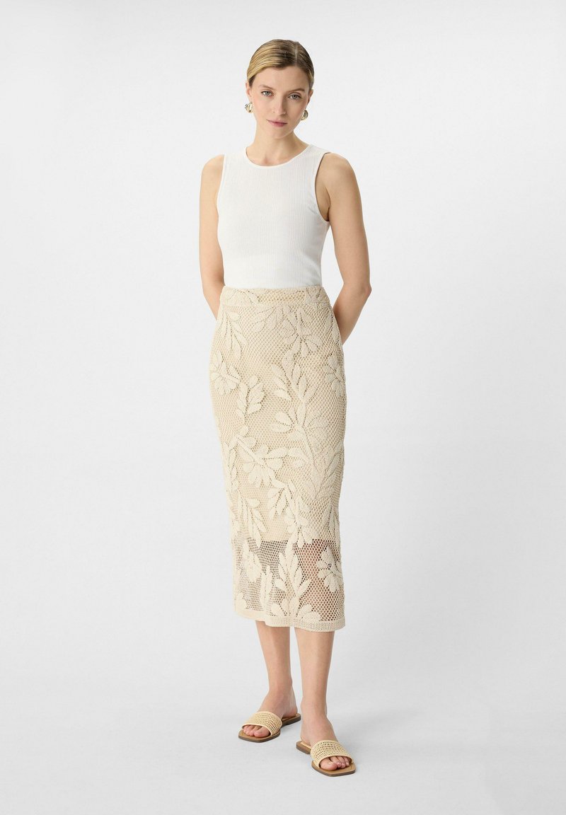 Beige Spitzen-Midi-Rock mit Blumenmustern; transparenter unterer Teil. Kombiniert mit einem weißen, figurbetonten Tanktop und geflochtenen Sandalen.