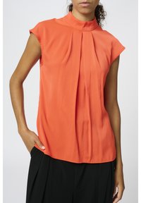 Blouse sans manches orange avec col montant, ornée de douces plis sur le devant, et réalisée dans un tissu lisse et léger.